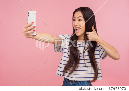 Portrait of satisfied friendly asian girl with brunette hair taking selfie and showing thumbs up gesture, communicating on video call, online chatting. indoor studio shot isolated on pink background Portrait of satisfied friendly asian girl with brunette hair taking selfie and showing thumbs up gesture, communicating on video call, online chatting. indoor studio shot isolated on pink background 87972070