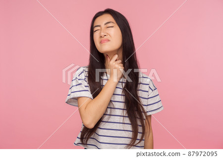 Sore throat. Ill girl with brunette hair in striped t-shirt touching neck, suffering thyroid disorder or tonsillitis, inflamed throat, medical concept. indoor studio shot isolated on pink background Sore throat. Ill girl with brunette hair in striped t-shirt touching neck, suffering thyroid disorder or tonsillitis, inflamed throat, medical concept. indoor studio shot isolated on pink background 87972095