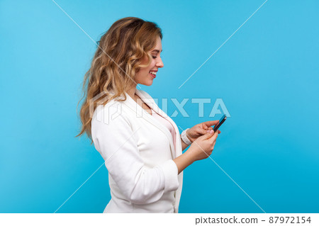 Side view of positive charming woman with wavy hair in white jacket typing message on cellphone and smiling, using mobile apps, online communication in social network. studio shot, blue background Side view of positive charming woman with wavy hair in white jacket typing message on cellphone and smiling, using mobile apps, online communication in social network. studio shot, blue background 87972154