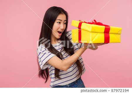 Satisfied with gift. Portrait of amazed girl with brunette hair in t-shirt peeking inside box and smiling excitedly, pleasantly surprised by present. indoor studio shot isolated on pink background 87972194