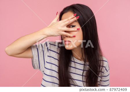 Portrait of nosy curious girl with brunette hair in striped t-shirt peeking through fingers, watching secrets, rumors, spying with inquisitive glance. indoor studio shot isolated on pink background 87972409
