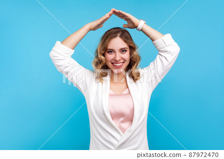 Life insurance. Portrait of cheerful woman with wavy hair and charming smile holding hands in gesture of house roof over head and smiling, feeling safe. indoor studio shot isolated on blue background 87972470