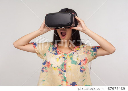Surprised woman wearing VR glasses and watching virtual world with amazed shocked expression, playing video game, experiencing strong emotions while using simulator. indoor studio shot gray background 87972489