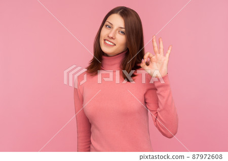 Everything is fine. Cheerful brunette woman in pink sweater showing ok gesture with hand looking at camera with toothy smile, approval. Indoor studio shot isolated on pink background Everything is fine. Cheerful brunette woman in pink sweater showing ok gesture with hand looking at camera with toothy smile, approval. Indoor studio shot isolated on pink background 87972608