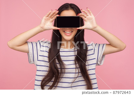 Anonymous mobile user. Portrait of girl with long brunette hair in striped t-shirt hiding eyes with cell phone and smiling, unknown internet customer. indoor studio shot isolated on pink background Anonymous mobile user. Portrait of girl with long brunette hair in striped t-shirt hiding eyes with cell phone and smiling, unknown internet customer. indoor studio shot isolated on pink background 87972643