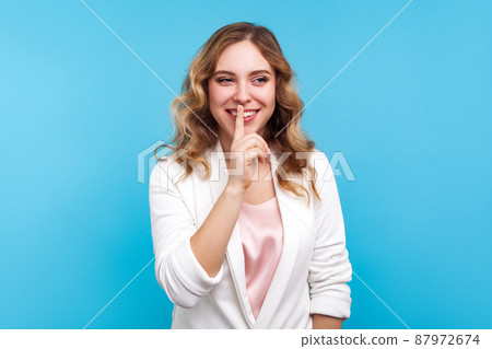 Please, be quiet, Portrait of positive lady with wavy hair in white jacket smiling and making hush sign with finger on lips, asking for silence and secrecy, keep calm. studio shot, blue background 87972674