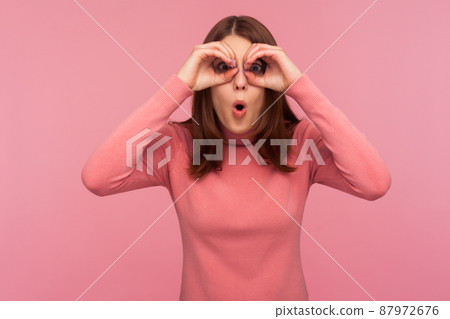 Funny curious woman with brown hair spying looking through holes in her hands, making binoculars, surprised with seen. Indoor studio shot isolated on pink background 87972676