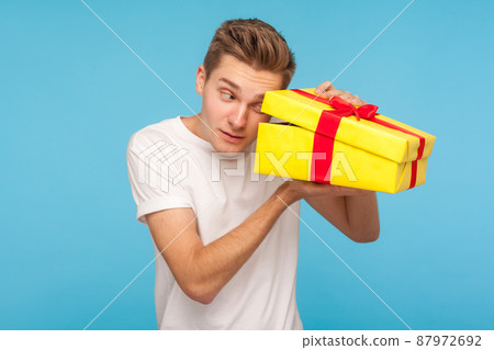 Nosy impatient man in white t-shirt looking inside gift box with curious happy expression, opening present before celebration, peeking with interest. indoor studio shot isolated on blue background 87972692
