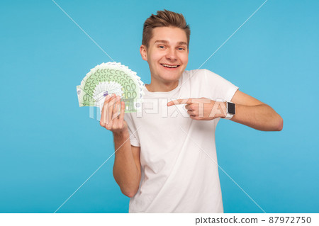 Overjoyed excited man in white t-shirt happy to hold big money, pointing to euro bills in hand and smiling excitedly, enjoying unexpected lottery win. indoor studio shot isolated on blue background Overjoyed excited man in white t-shirt happy to hold big money, pointing to euro bills in hand and smiling excitedly, enjoying unexpected lottery win. indoor studio shot isolated on blue background 87972750