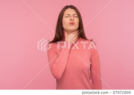 Sad woman with brown hair palping neck feeling throat ache and pain during swallow, tonsillitis inflammation. Indoor studio shot isolated on pink background Sad woman with brown hair palping neck feeling throat ache and pain during swallow, tonsillitis inflammation. Indoor studio shot isolated on pink background 87972809