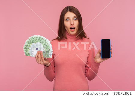 Extremely surprised brunette woman in pink sweater holding smartphone with empty screen and fan of euros looking at camera with shocked expression. Indoor studio shot isolated on pink background 87972901