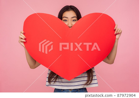 Portrait of playful charming girl peeking out of huge red heart, looking with alluring eyes at camera and holding symbol of love, romantic emotions. indoor studio shot isolated on pink background Portrait of playful charming girl peeking out of huge red heart, looking with alluring eyes at camera and holding symbol of love, romantic emotions. indoor studio shot isolated on pink background 87972904