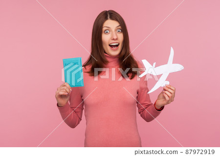 Shocked woman with brown hair in pink sweater holding in hands paper plane and passport, preparing for sudden journey, amazed with adventure. Indoor studio shot isolated on pink background Shocked woman with brown hair in pink sweater holding in hands paper plane and passport, preparing for sudden journey, amazed with adventure. Indoor studio shot isolated on pink background 87972919
