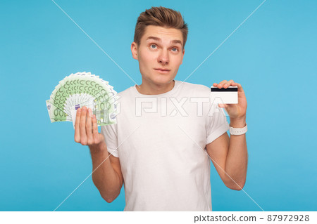 Pensive man in white t-shirt holding euro bills and credit card, thinking over benefits of bank loan, consumer pondering deposit percentage, doubting. indoor studio shot isolated on blue background 87972928