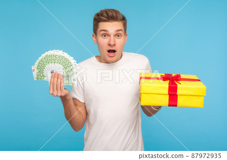 Wow, bonuses on holiday shopping. Amazed shocked man in white t-shirt holding gift box and euro banknotes, surprised by present and big cashback, showing money. studio shot isolated on blue background 87972935
