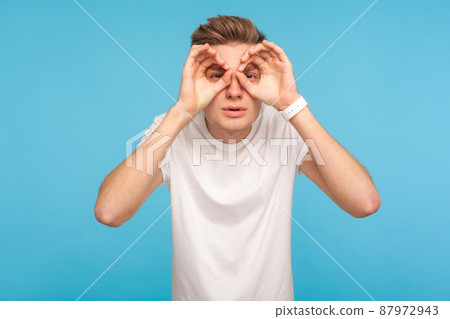 Portrait of curious man in casual white t-shirt looking through fingers in binoculars gesture, observing distant with attentive look, watching afar. indoor studio shot isolated on blue background 87972943