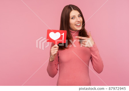 Smiling brunette woman in pink sweater pointing finger at sign with like counter, blogger waiting for feedback and positive rate. Indoor studio shot isolated on pink background Smiling brunette woman in pink sweater pointing finger at sign with like counter, blogger waiting for feedback and positive rate. Indoor studio shot isolated on pink background 87972945