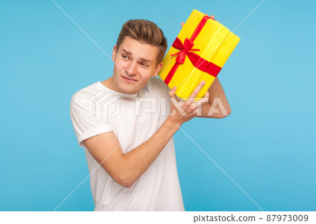 Young man in white t-shirt holding birthday box near ear, listening what's inside and expressing disbelief doubts about present, dissatisfied with gift. indoor studio shot isolated on blue background Young man in white t-shirt holding birthday box near ear, listening what's inside and expressing disbelief doubts about present, dissatisfied with gift. indoor studio shot isolated on blue background 87973009