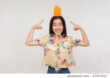 Dream to buy house. Happy asian girl with brunette hair in blouse pointing at paper building on head and smiling contentedly, hoping for property loan. indoor studio shot isolated on gray background Dream to buy house. Happy asian girl with brunette hair in blouse pointing at paper building on head and smiling contentedly, hoping for property loan. indoor studio shot isolated on gray background 87973027