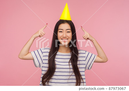Portrait of joyful funny asian girl pointing at party cone on head and looking at camera with toothy smile, enjoying birthday party, holiday celebration. indoor studio shot isolated on pink background Portrait of joyful funny asian girl pointing at party cone on head and looking at camera with toothy smile, enjoying birthday party, holiday celebration. indoor studio shot isolated on pink background 87973067