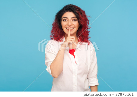 Shh, keep quiet. Portrait of positive hipster woman with fancy red hair in white shirt holding finger on lips, hushing and showing silence gesture. indoor studio shot isolated on blue background Shh, keep quiet. Portrait of positive hipster woman with fancy red hair in white shirt holding finger on lips, hushing and showing silence gesture. indoor studio shot isolated on blue background 87973161