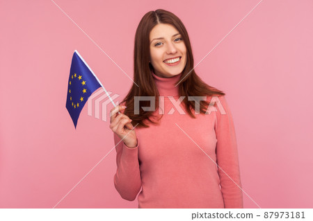 Happy patriotic woman with brown hair holding eu flag in hand looking at camera with toothy smile, patriotism, flag day celebration. Indoor studio shot isolated on pink background Happy patriotic woman with brown hair holding eu flag in hand looking at camera with toothy smile, patriotism, flag day celebration. Indoor studio shot isolated on pink background 87973181