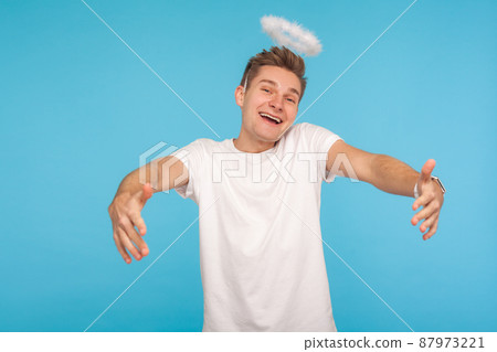 Let me embrace you. Angelic kind-hearted man with halo above head outstretching hands to hug and looking at camera with childish joyful expression. indoor studio shot isolated on blue background 87973221