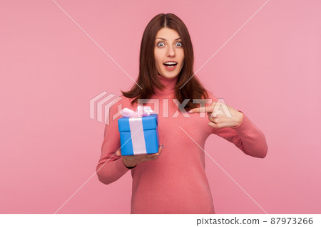 Excited surprised brunette woman in pink sweater pointing finger at blue giftbox in her hand looking at camera with wondered expression. Indoor studio shot isolated on pink background 87973266