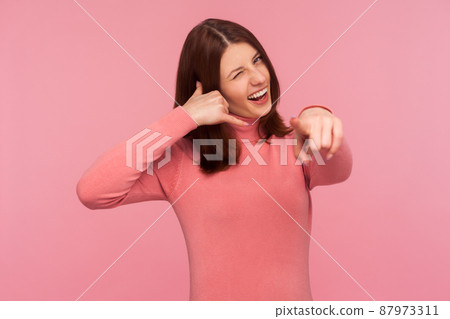Communicative positive brunette woman showing call gesture and pointing finger at you, waiting for your call. Indoor studio shot isolated on pink background Communicative positive brunette woman showing call gesture and pointing finger at you, waiting for your call. Indoor studio shot isolated on pink background 87973311