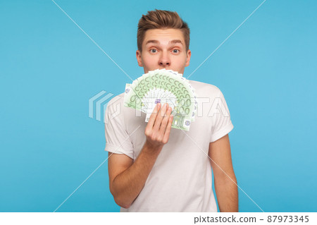 Portrait of young astonished man in casual white t-shirt holding euro bills, hiding half face and peeking out of money with surprised shocked eyes. indoor studio shot isolated on blue background Portrait of young astonished man in casual white t-shirt holding euro bills, hiding half face and peeking out of money with surprised shocked eyes. indoor studio shot isolated on blue background 87973345