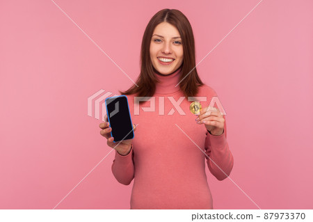 Bitcoin cryptocurrency payments. Happy excited woman with brown hair holding btc golden coin and cellphone looking at camera with toothy smile. Indoor studio shot isolated on pink background Bitcoin cryptocurrency payments. Happy excited woman with brown hair holding btc golden coin and cellphone looking at camera with toothy smile. Indoor studio shot isolated on pink background 87973370