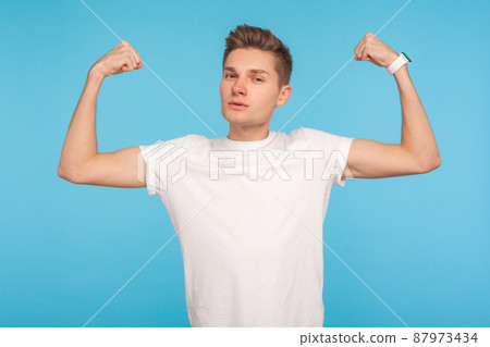 I'm strong. Portrait of slim young man in casual white t-shirt raising hands to show biceps and looking confident at camera, feeling power and strength. indoor studio shot isolated on blue background 87973434