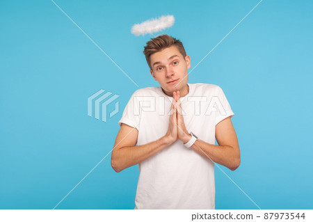 Portrait of angelic nice-looking man with halo above head holding hands in prayer and looking at camera with timid shy expression, obedient behavior. indoor studio shot isolated on blue background Portrait of angelic nice-looking man with halo above head holding hands in prayer and looking at camera with timid shy expression, obedient behavior. indoor studio shot isolated on blue background 87973544