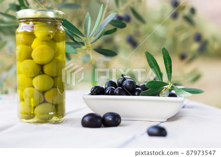 canned olives in a glass jar, oil and olives on the table against the background of the olive garden canned olives in a glass jar, oil and olives on the table against the background of the olive garden 87973547