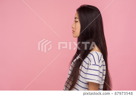 Side view of serious calm beautiful asian girl with long brunette hair in striped t-shirt looking at copy space with concentrated thoughtful expression. indoor studio shot isolated on pink background Side view of serious calm beautiful asian girl with long brunette hair in striped t-shirt looking at copy space with concentrated thoughtful expression. indoor studio shot isolated on pink background 87973548