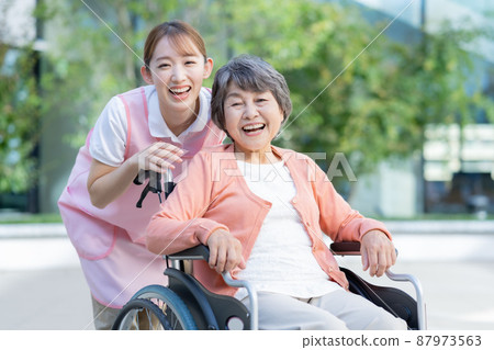 Portrait of a senior woman in a wheelchair and a young female nurse pushing a wheelchair. 87973563