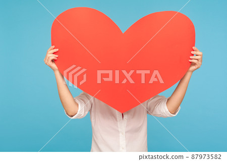 Anonymous romantic dating on Valentine's Day. Unknown woman hiding behind large heart, expressing her great romantic feelings with big symbol of love. indoor studio shot isolated on blue background 87973582