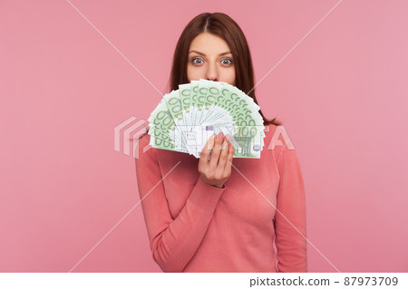 Amazed happy woman with brown hair in pink sweater hiding face behind fan of euro banknotes, interest-free cash withdrawal. Indoor studio shot isolated on pink background Amazed happy woman with brown hair in pink sweater hiding face behind fan of euro banknotes, interest-free cash withdrawal. Indoor studio shot isolated on pink background 87973709