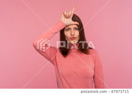 Unhappy disappointed woman with brown hair in pink sweater holding fingers near head showing loser gesture, frustration, misfortune. Indoor studio shot isolated on pink background Unhappy disappointed woman with brown hair in pink sweater holding fingers near head showing loser gesture, frustration, misfortune. Indoor studio shot isolated on pink background 87973715