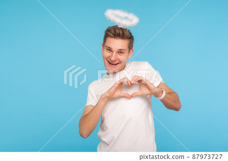 Portrait of happy angelic man with halo above head making heart shape with hands and smiling to camera, sharing romantic amorous feelings, declaration of love. studio shot isolated on blue background Portrait of happy angelic man with halo above head making heart shape with hands and smiling to camera, sharing romantic amorous feelings, declaration of love. studio shot isolated on blue background 87973727