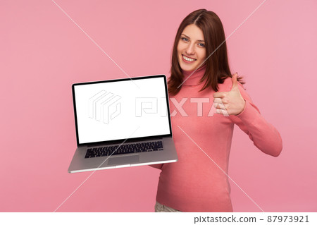 Excited satisfied woman with brown hair in pink sweater holding laptop with empty blank display and showing thumbs up, pleased with online application. Indoor studio shot isolated on pink background 87973921