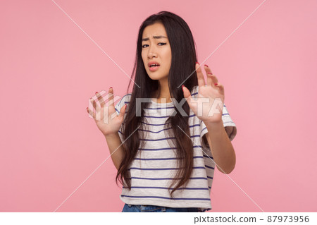 I'm scared, Portrait of panicking girl with long brunette hair in striped t-shirt expressing fear, holding hands up, looking terrified as noticing something terrible. indoor studio shot isolated I'm scared, Portrait of panicking girl with long brunette hair in striped t-shirt expressing fear, holding hands up, looking terrified as noticing something terrible. indoor studio shot isolated 87973956