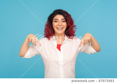 Advertising area. Happy cheerful hipster woman with fancy red hair in white shirt pointing down, showing place for commercial, recommending to subscribe. indoor studio shot isolated on blue background 87973983
