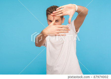 Portrait of curious man in white t-shirt gesturing picture frame with hands, looking through fingers and focusing on interesting moment, taking photo. indoor studio shot isolated on blue background 87974002