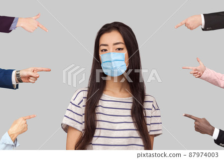 Many different people hands are pointing on guilty alone young woman with surgical medical mask and blame her. she looking at camera with sad face. indoor studio shot, isolated on gray background. Many different people hands are pointing on guilty alone young woman with surgical medical mask and blame her. she looking at camera with sad face. indoor studio shot, isolated on gray background. 87974003