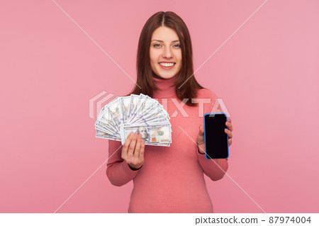 Cheerful brunette woman in pink sweater holding dollars banknotes and cell phone with mock up blank display, online payments, cashback. Indoor studio shot isolated on pink background Cheerful brunette woman in pink sweater holding dollars banknotes and cell phone with mock up blank display, online payments, cashback. Indoor studio shot isolated on pink background 87974004