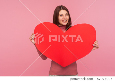 Happy cheerful brunette woman holding in hands big red paper heart and smiling at camera, love and romance. Indoor studio shot isolated on pink background 87974007