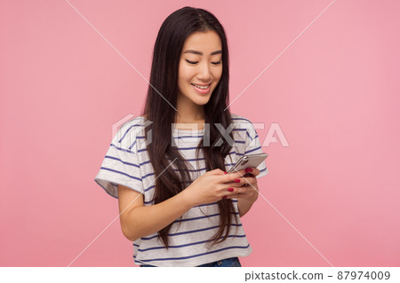 Portrait of happy girl with brunette hair in t-shirt typing message on mobile phone and smiling, reading funny post in social network, chatting online. indoor studio shot isolated on pink background Portrait of happy girl with brunette hair in t-shirt typing message on mobile phone and smiling, reading funny post in social network, chatting online. indoor studio shot isolated on pink background 87974009