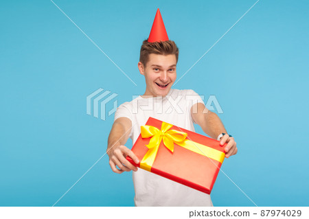 Surprise. Present for you. Funny glad generous man in white t-shirt and party cone on head giving gift box and smiling with childish carefree expression. indoor studio shot isolated on blue background 87974029