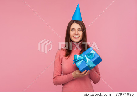 Optimistic positive brunette woman in cone hat on head holding gift with bow and looking at camera with toothy smile, gifts and bonuses, celebration. Indoor studio shot isolated on pink background 87974080